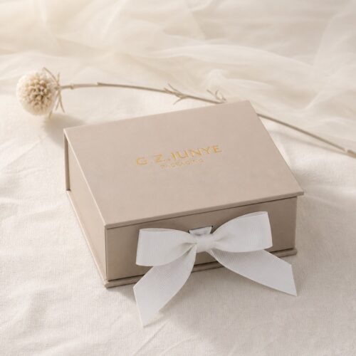 Beige flip jewelry box with a white bow styled on cream fabric beside dried flowers