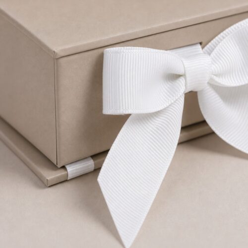 White grosgrain ribbon bow at the front of a beige jewelry box, with the ribbon emerging from under the base