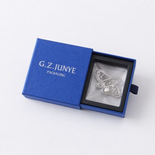 Blue square drawer jewelry box with the tray drawn out, holding a brooch in the black blister frame and clear bag